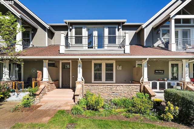 view of front facade featuring a balcony, stone siding, a porch, and roof with shingles