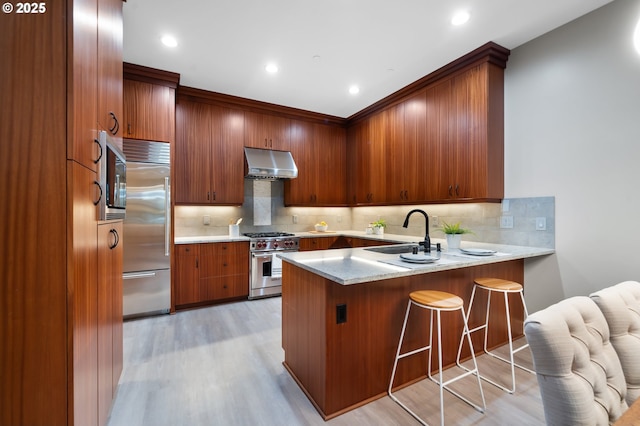 kitchen featuring a breakfast bar, built in appliances, a peninsula, under cabinet range hood, and a sink