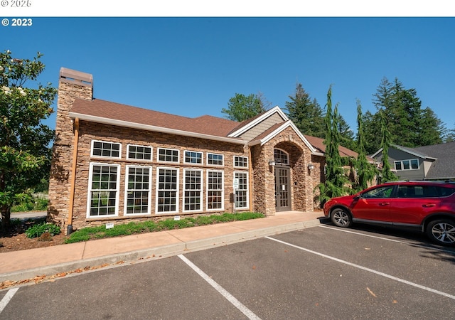 view of front facade with uncovered parking, a chimney, stone siding, and roof with shingles