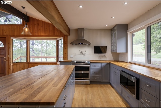 kitchen with wood counters, wall chimney range hood, gray cabinets, appliances with stainless steel finishes, and light wood finished floors