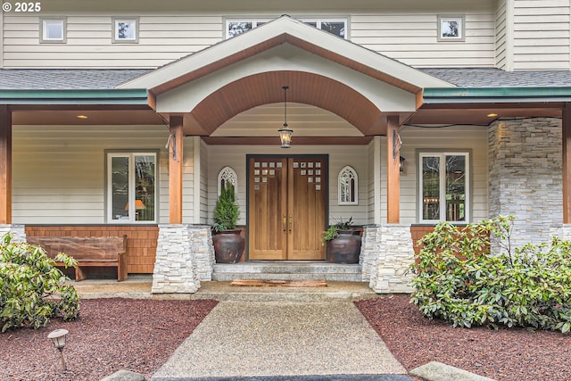 doorway to property featuring stone siding, a porch, and roof with shingles