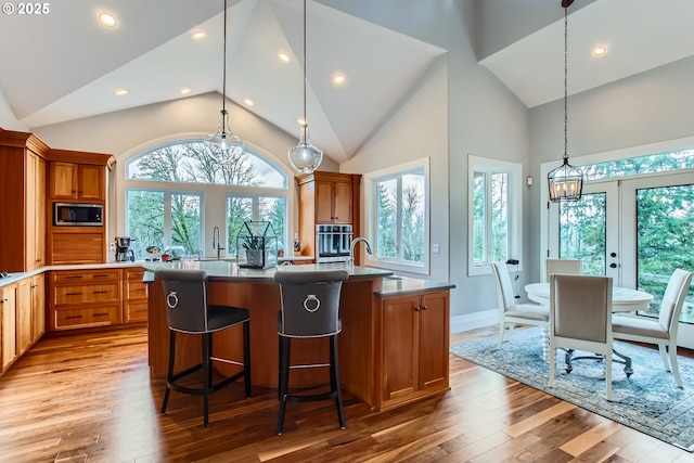 kitchen featuring a center island with sink, brown cabinetry, appliances with stainless steel finishes, wood finished floors, and hanging light fixtures