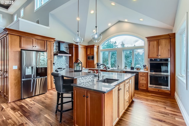 kitchen featuring a center island with sink, stainless steel appliances, dark countertops, a sink, and wall chimney exhaust hood