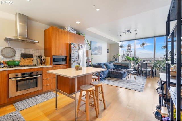 kitchen with light wood finished floors, wall chimney exhaust hood, stainless steel appliances, light countertops, and tasteful backsplash