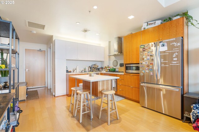 kitchen featuring light wood-type flooring, wall chimney exhaust hood, light countertops, a center island, and appliances with stainless steel finishes