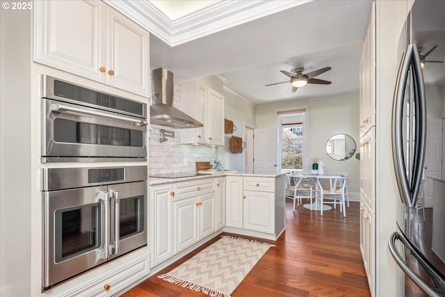 kitchen with crown molding, ceiling fan, stainless steel appliances, wall chimney exhaust hood, and dark wood-style flooring