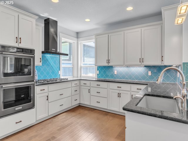 14509 SE Faircrest St, Clackamas, OR 97015 - kitchen featuring appliances with stainless steel finishes, wall chimney range hood, light wood finished floors, dark stone countertops, and white cabinetry