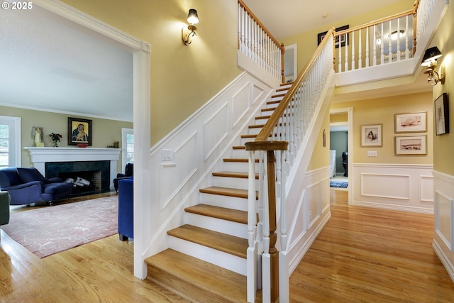 staircase featuring a decorative wall, a fireplace, wood finished floors, ornamental molding, and wainscoting