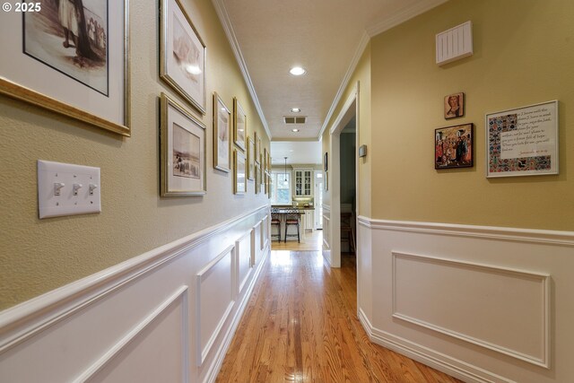 hallway with light wood-style flooring, a wainscoted wall, crown molding, and visible vents