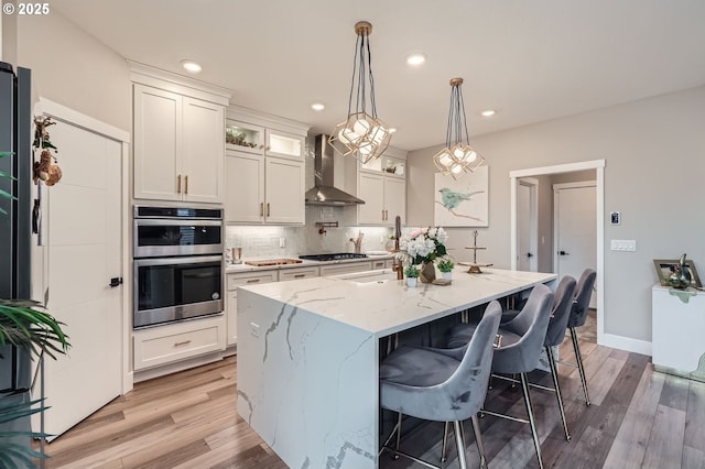 kitchen featuring decorative backsplash, double oven, light wood-style flooring, and wall chimney range hood