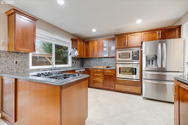 kitchen featuring dark stone countertops, stainless steel appliances, decorative backsplash, glass insert cabinets, and brown cabinets