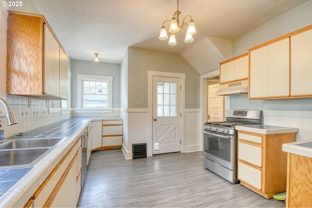 kitchen with appliances with stainless steel finishes, a wainscoted wall, under cabinet range hood, light wood-style flooring, and tile countertops