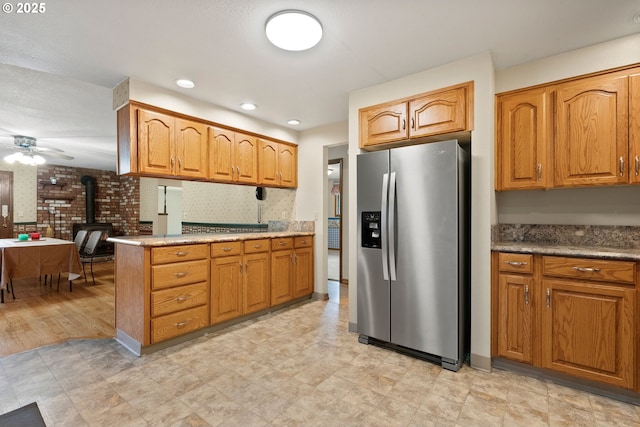 kitchen with stainless steel fridge, a wood stove, a ceiling fan, light flooring, and brown cabinets