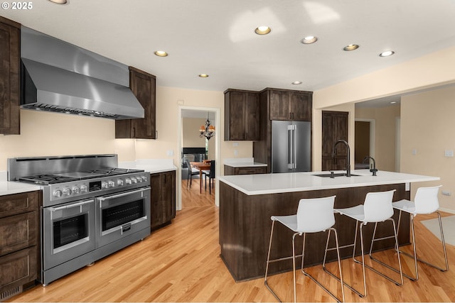 kitchen featuring an island with sink, high quality appliances, light countertops, light wood-type flooring, and wall chimney range hood