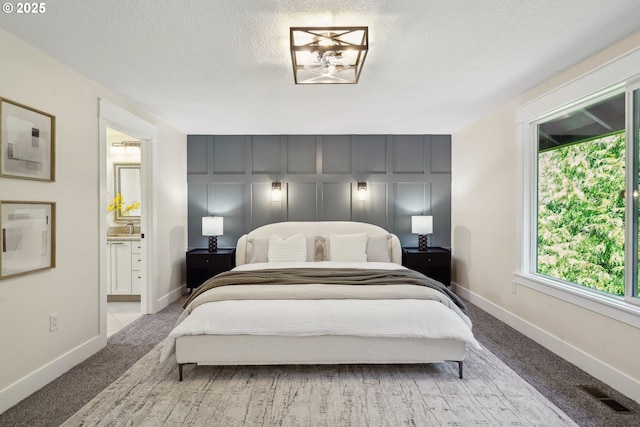 carpeted bedroom featuring visible vents, a decorative wall, a textured ceiling, ensuite bath, and baseboards