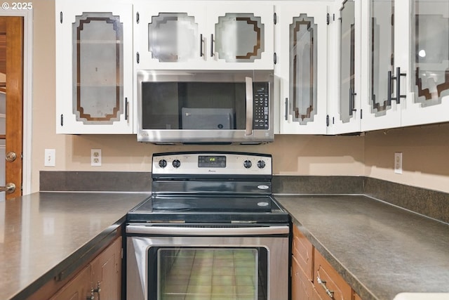 kitchen with brown cabinetry, white cabinets, glass insert cabinets, appliances with stainless steel finishes, and dark countertops