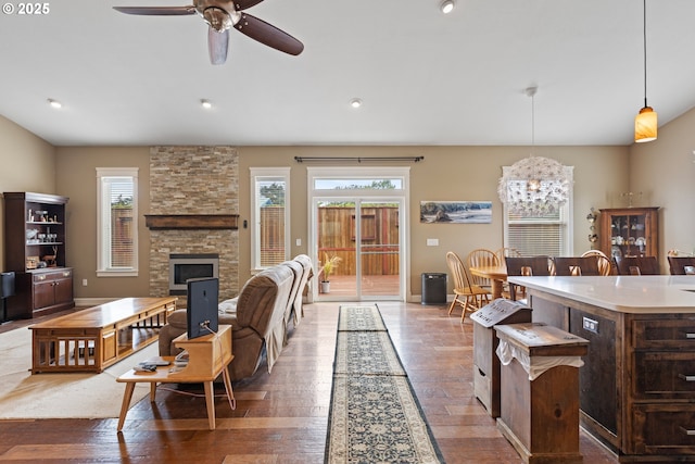 living area with wood-type flooring, a stone fireplace, a chandelier, ceiling fan, and baseboards
