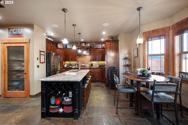 kitchen with decorative backsplash, freestanding refrigerator, stone finish flooring, under cabinet range hood, and a sink