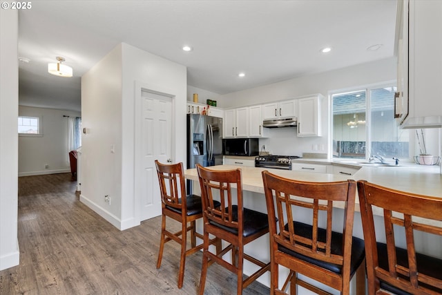 kitchen with a peninsula, under cabinet range hood, white cabinetry, wood finished floors, and stainless steel appliances