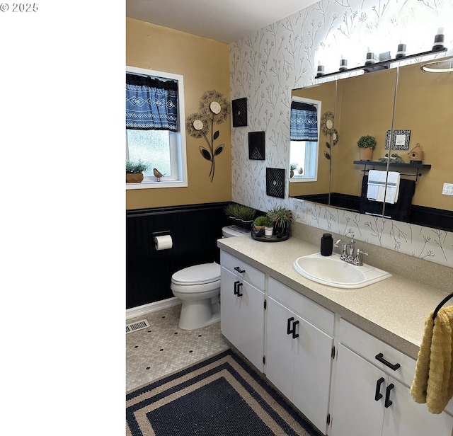 bathroom with toilet, vanity, visible vents, wainscoting, and wallpapered walls