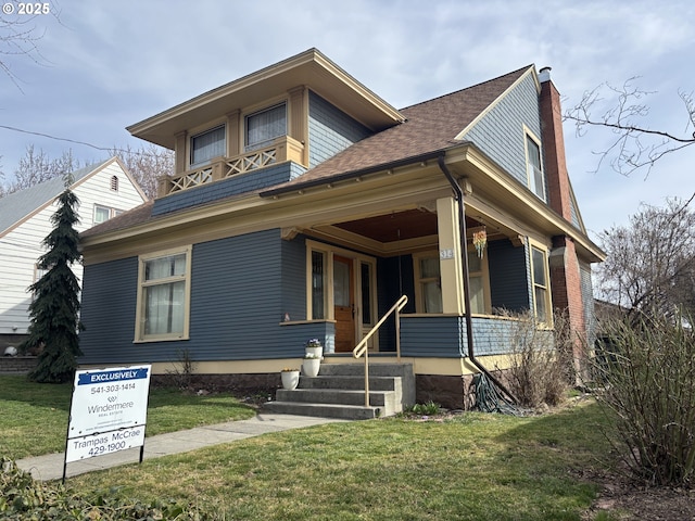 view of front facade featuring a chimney, covered porch, a front yard, and a shingled roof