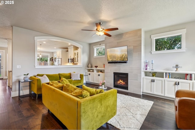 living room featuring dark wood-type flooring, baseboards, ceiling fan, a tiled fireplace, and a textured ceiling