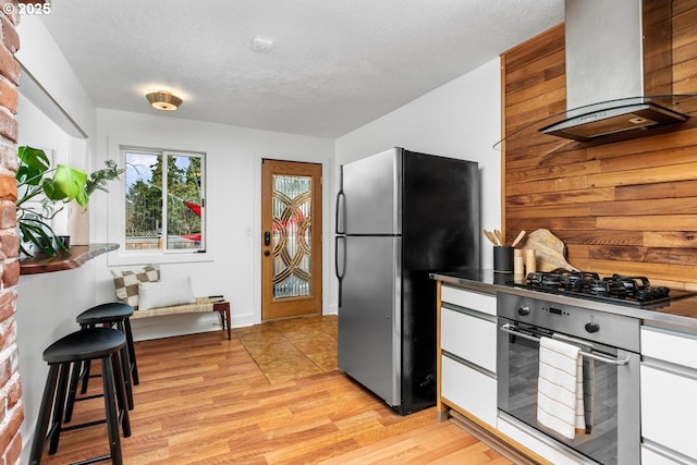 kitchen featuring dark countertops, a textured ceiling, light wood-style floors, appliances with stainless steel finishes, and wall chimney exhaust hood