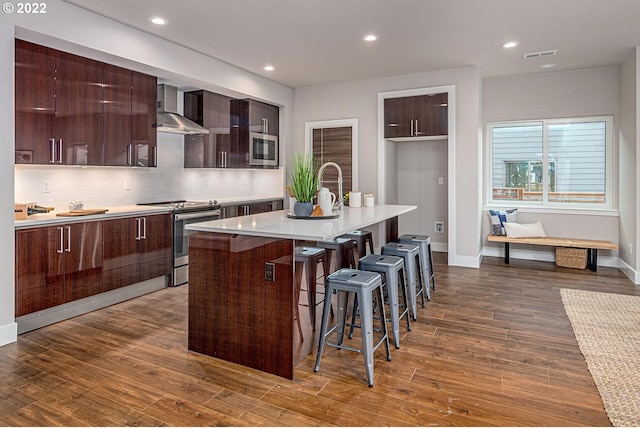 kitchen with wood-type flooring, stainless steel stove, a center island with sink, and wall chimney exhaust hood