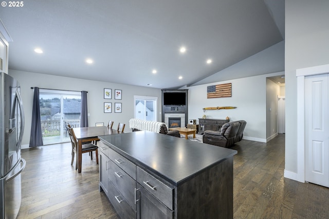 kitchen featuring stainless steel refrigerator with ice dispenser, dark countertops, a glass covered fireplace, dark wood finished floors, and a kitchen island