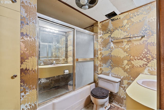 bathroom with bath / shower combo with glass door, vanity, toilet, and wallpapered walls