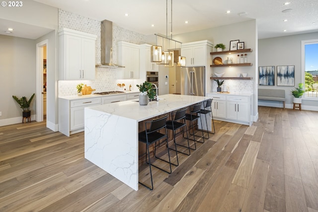 kitchen featuring appliances with stainless steel finishes, an island with sink, light hardwood / wood-style floors, wall chimney exhaust hood, and white cabinets