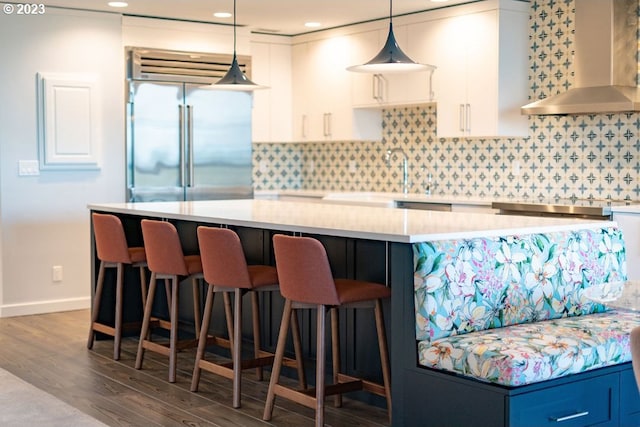 kitchen featuring hanging light fixtures, wall chimney exhaust hood, stainless steel built in refrigerator, white cabinetry, and dark hardwood / wood-style floors