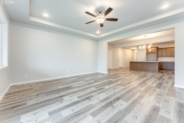 unfurnished living room featuring ceiling fan with notable chandelier, light wood-type flooring, and a tray ceiling