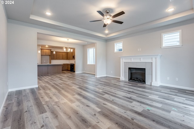 unfurnished living room featuring a tile fireplace, a raised ceiling, ceiling fan with notable chandelier, and light hardwood / wood-style flooring