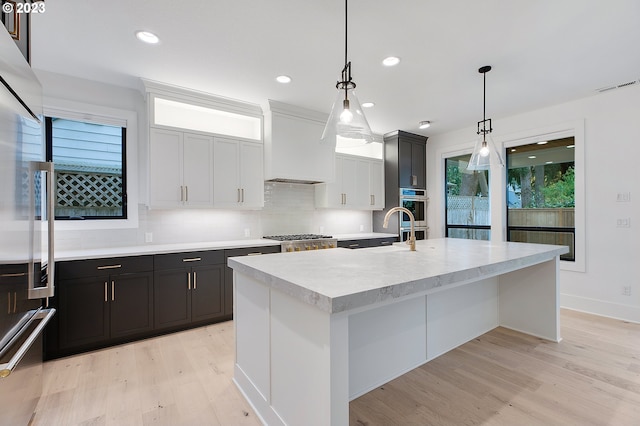 kitchen with light hardwood / wood-style flooring, backsplash, hanging light fixtures, a kitchen island with sink, and premium range hood