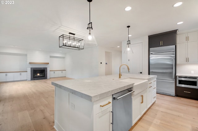 kitchen with a large fireplace, a center island with sink, decorative light fixtures, light wood-type flooring, and appliances with stainless steel finishes