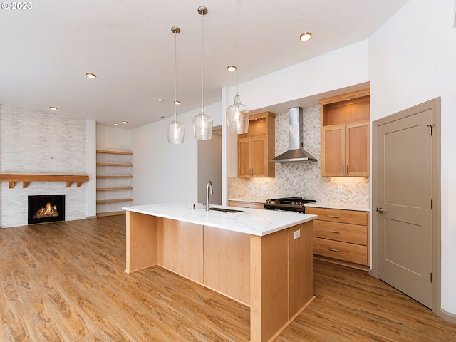 kitchen featuring a fireplace, an island with sink, light wood-type flooring, wall chimney range hood, and tasteful backsplash