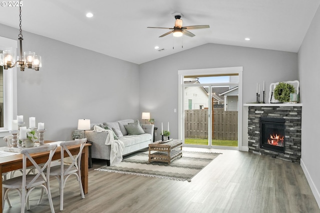 living room featuring wood-type flooring, vaulted ceiling, ceiling fan with notable chandelier, and a stone fireplace