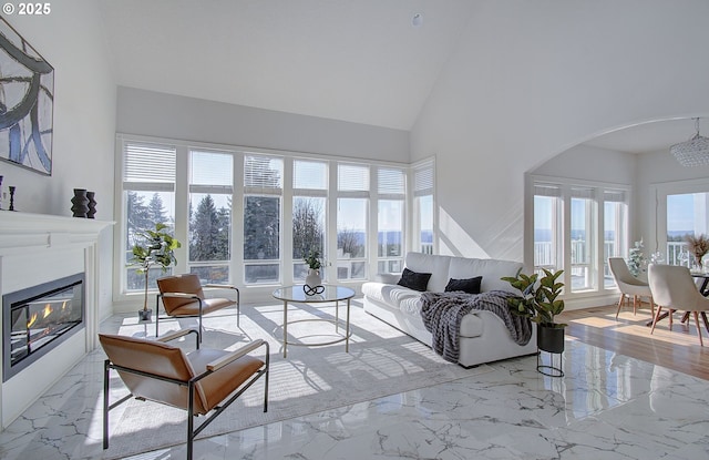 living room featuring arched walkways, a glass covered fireplace, marble finish floor, and high vaulted ceiling