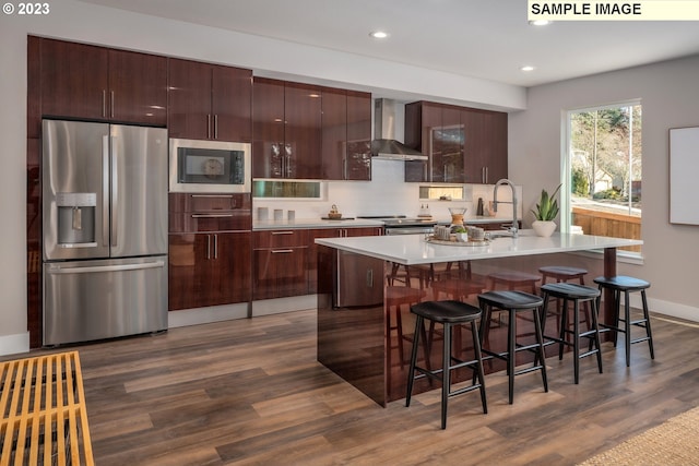 kitchen with stainless steel appliances, a kitchen breakfast bar, a center island with sink, dark hardwood / wood-style flooring, and wall chimney range hood