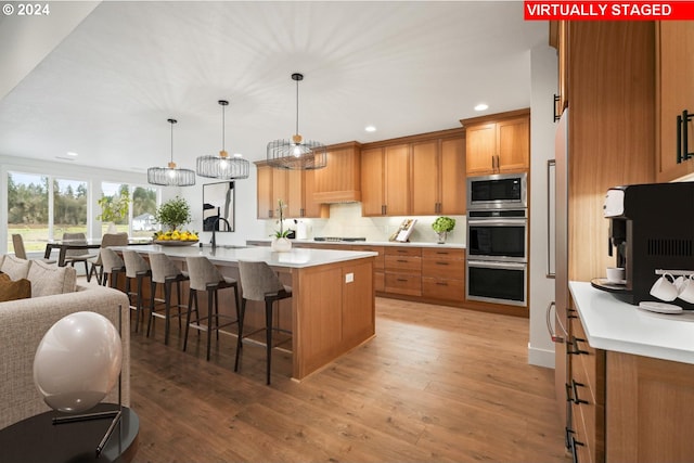 kitchen featuring decorative light fixtures, backsplash, stainless steel appliances, light hardwood / wood-style floors, and a center island with sink