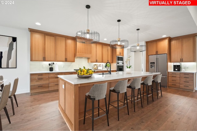 kitchen with decorative light fixtures, stainless steel appliances, light hardwood / wood-style floors, a center island with sink, and tasteful backsplash