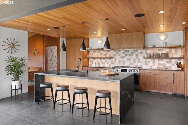 kitchen with decorative light fixtures, a breakfast bar area, an island with sink, backsplash, and stainless steel stove