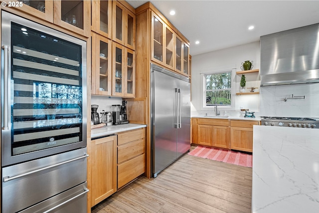 kitchen with built in refrigerator, wall chimney range hood, a sink, light wood finished floors, and light stone countertops