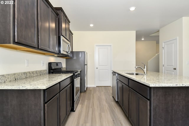 kitchen with stainless steel appliances, sink, light hardwood / wood-style flooring, light stone countertops, and a center island with sink