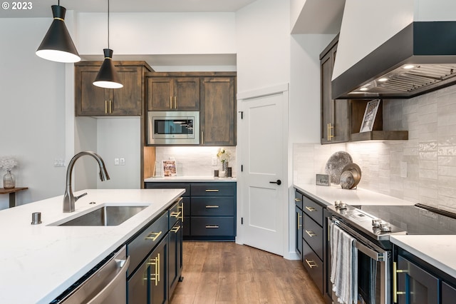 kitchen featuring dark wood-type flooring, a sink, hanging light fixtures, wall chimney range hood, and appliances with stainless steel finishes