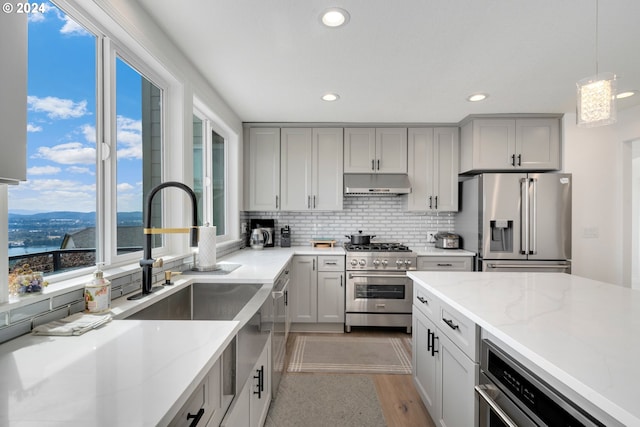 kitchen with pendant lighting, under cabinet range hood, light stone counters, backsplash, and appliances with stainless steel finishes