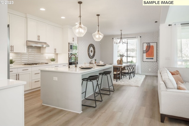 kitchen with appliances with stainless steel finishes, pendant lighting, white cabinets, a kitchen island with sink, and light hardwood / wood-style flooring