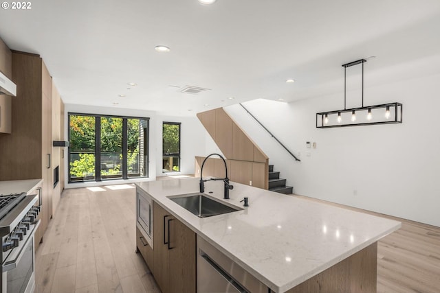 kitchen with appliances with stainless steel finishes, hanging light fixtures, light stone counters, light hardwood / wood-style floors, and a center island with sink