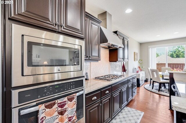 10703 NW Glenmore Way, Portland, OR 97229 - kitchen with appliances with stainless steel finishes, wall chimney exhaust hood, tasteful backsplash, light wood-style flooring, and light stone counters